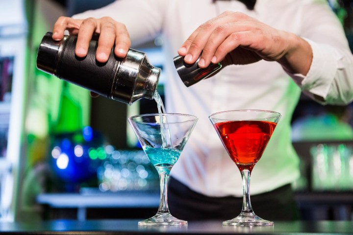 Bartender pouring blue and red cocktails into glasses with shaker, background blurred.