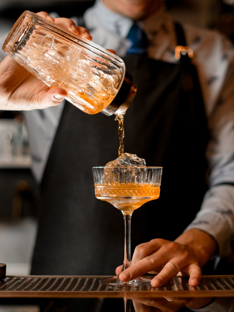 Bartender pouring cocktail from shaker into glass with ice cube on bar counter.